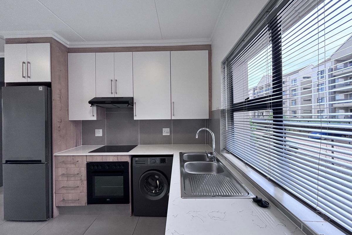 Modern kitchen featuring white and wood-grain cabinetry, stone countertops, stainless steel sink, built-in oven, hob, and extractor fan with large window and venetian blinds.