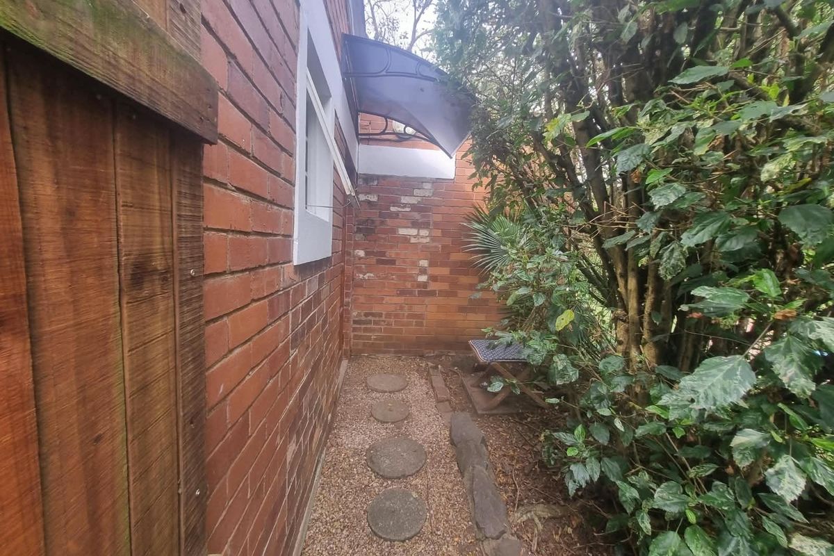 A narrow side passage featuring a red brick wall, a small protective awning over a window, and a gravel path with stone pavers.