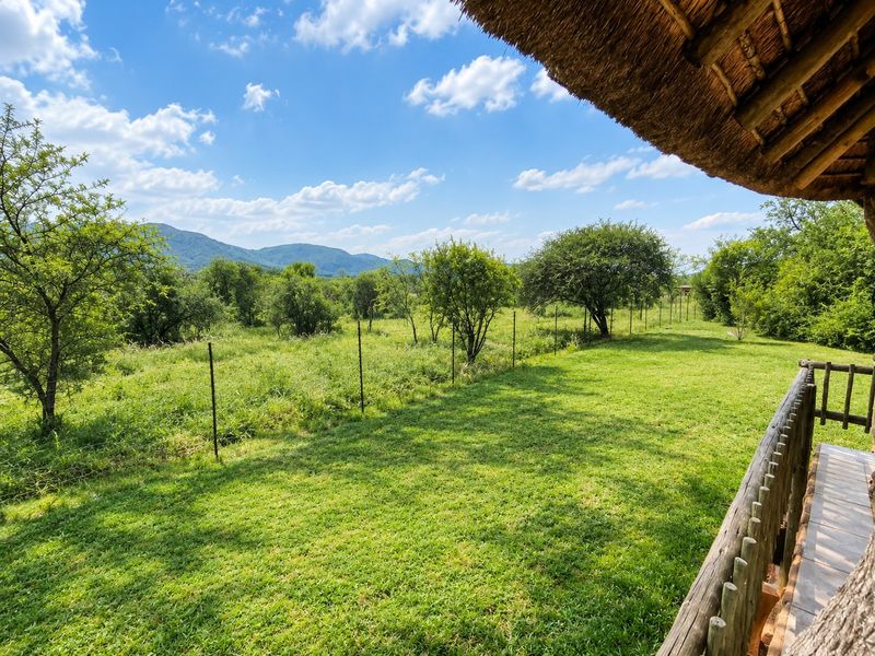 A covered patio area with a traditional thatch roof and wooden railing, overlooking a lush green garden and mountain landscape.