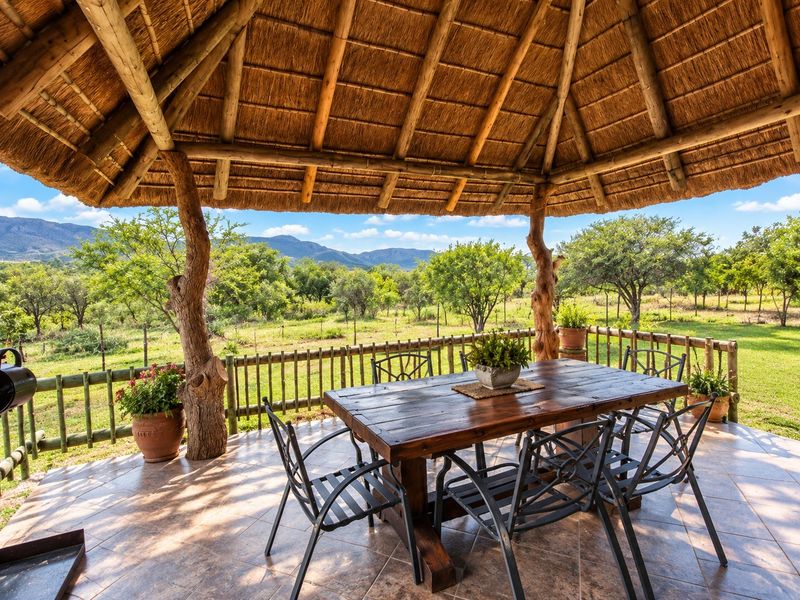 A covered outdoor patio featuring a traditional thatched roof, timber support columns, and tiled flooring, overlooking a scenic rural landscape.
