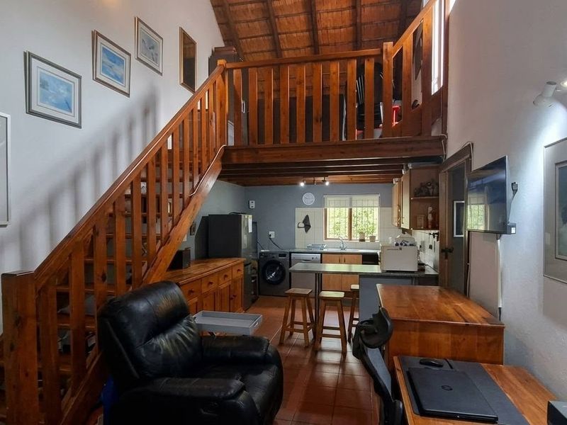 Open-plan living area featuring a wooden staircase leading to a mezzanine level, exposed timber ceiling, and a functional kitchen space with tiled flooring.