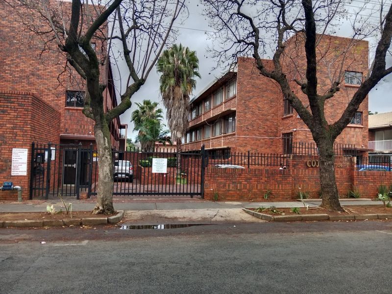 Street view of a multi-story face brick apartment complex featuring secure perimeter fencing, gated vehicle access, and established trees.