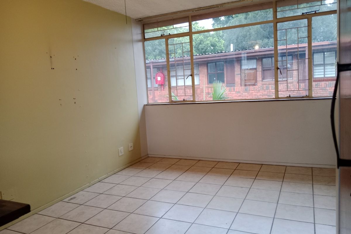 An empty living room featuring tiled flooring and large windows, providing natural light and a view of the surrounding apartment complex.