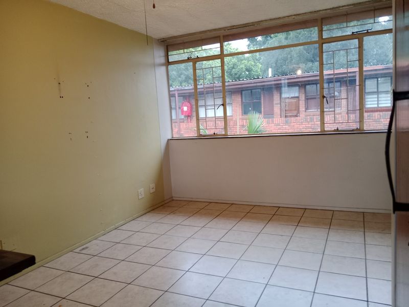 An empty living room featuring tiled flooring and large windows, providing natural light and a view of the surrounding apartment complex.