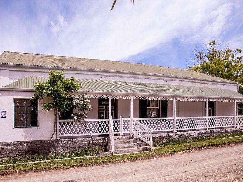 A charming historic cottage featuring a classic veranda with lattice detailing, a corrugated iron roof, and a stone foundation, set along a quiet road.