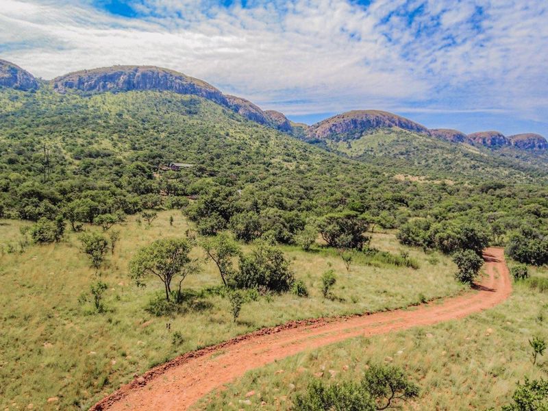 An aerial view of a vacant residential plot featuring a dirt access road, natural bushveld vegetation, and scenic mountain views.