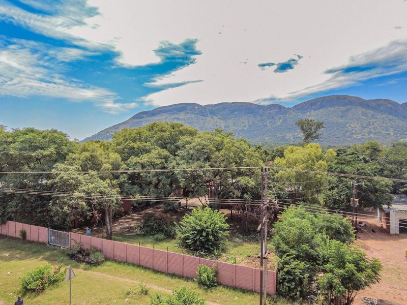 An aerial view of a residential plot featuring mature trees, a perimeter wall, and mountain views in the background.