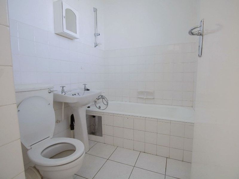 A functional bathroom featuring a pedestal basin, toilet, and bathtub with tiled finishes. The space requires minor repairs to the bath panel and grout.