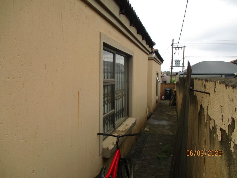 A narrow side passage of the house featuring a concrete walkway, security-fitted windows, and a boundary wall showing signs of weathering and paint deterioration.