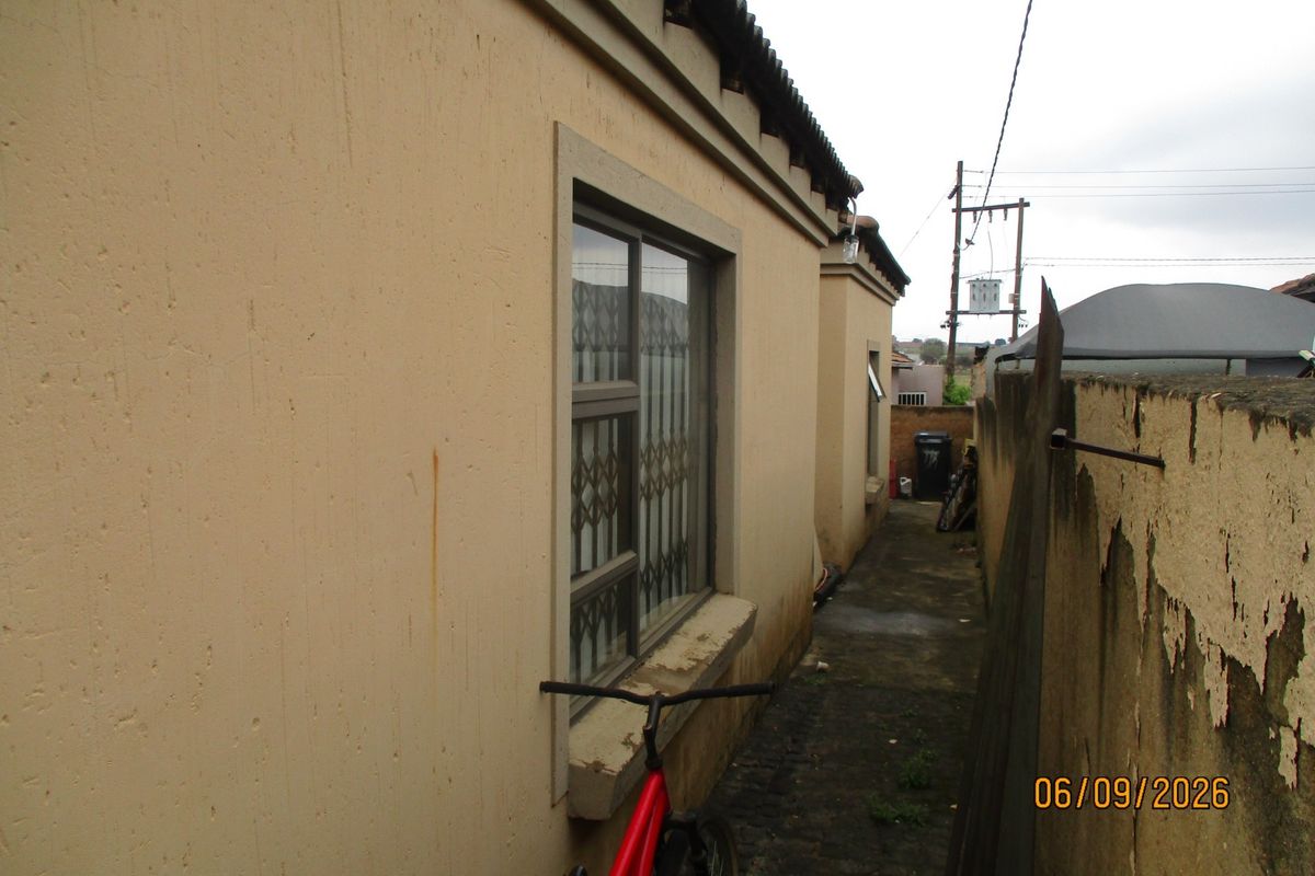 A narrow side passage of the house featuring a concrete walkway, security-fitted windows, and a boundary wall showing signs of weathering and paint deterioration.