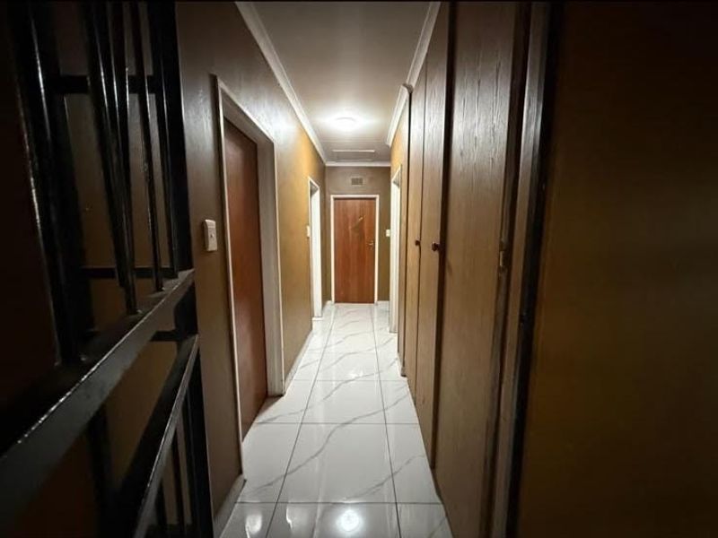 A long hallway featuring white marble-look tiled flooring, built-in wooden cupboards, and multiple interior doors leading to various rooms.