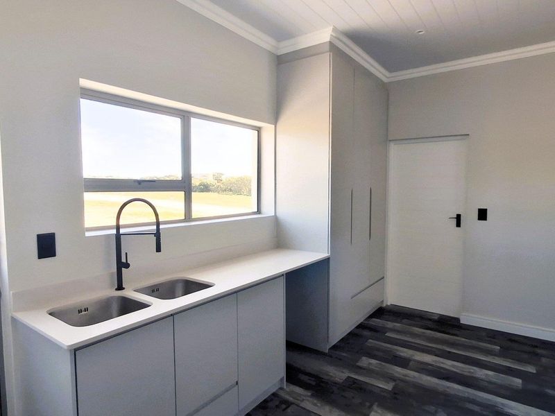 A modern, clean scullery featuring a double basin sink, stone countertops, ample built-in cabinetry, and durable laminate flooring with a view of the outdoors.