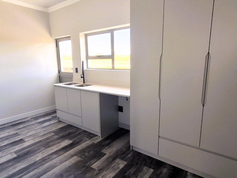 A modern, minimalist kitchen featuring sleek white cabinetry, a clean countertop with a sink, and contemporary wood-look laminate flooring.