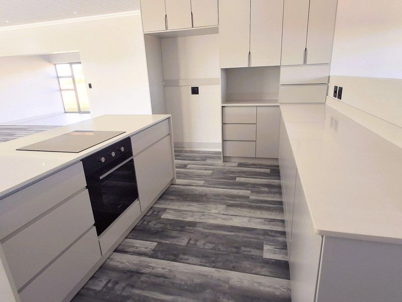 Modern, minimalist kitchen featuring sleek white cabinetry, stone countertops, a built-in oven, and a central island with an integrated cooktop.
