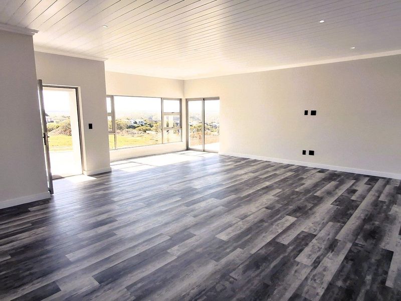 A spacious, light-filled living area featuring modern laminate flooring, a white tongue and groove ceiling, and large aluminium windows with scenic views.