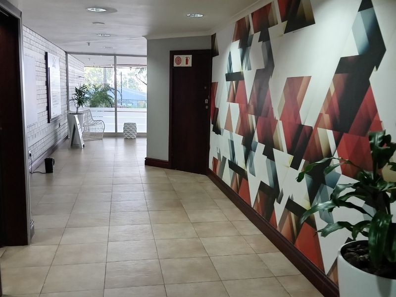 A spacious office hallway featuring ceramic floor tiles, recessed lighting, and a decorative geometric wall mural leading toward a glass-fronted entrance.