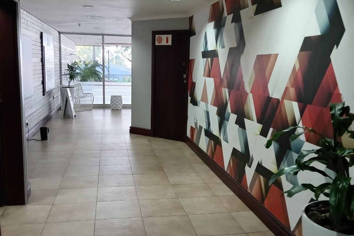 A spacious office hallway featuring ceramic floor tiles, recessed lighting, and a decorative geometric wall mural leading toward a glass-fronted entrance.