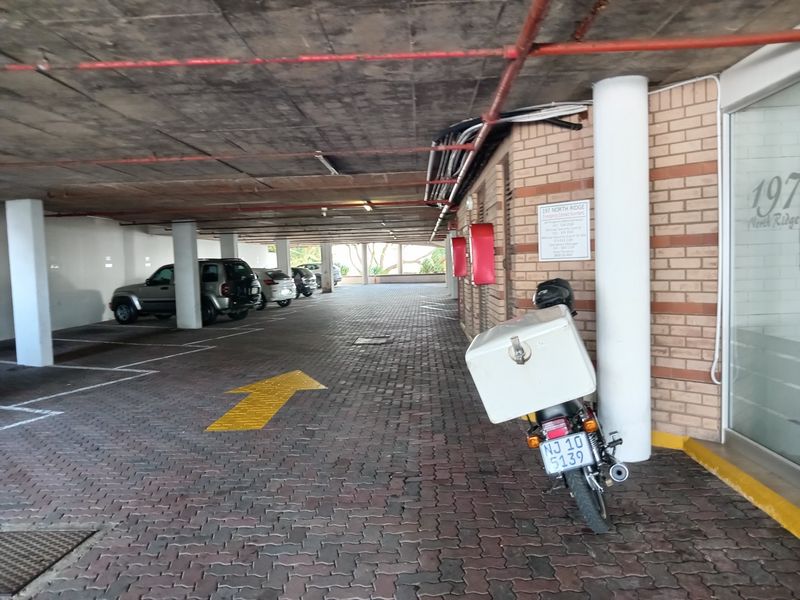 Covered parking garage area featuring paved flooring, exposed concrete ceiling with fire suppression systems, and brick wall detailing.