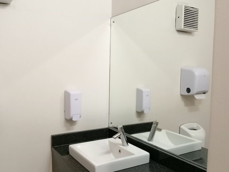 A clean, modern office restroom vanity area featuring a granite countertop, integrated ceramic basin, and wall-mounted mirror with essential utility dispensers.