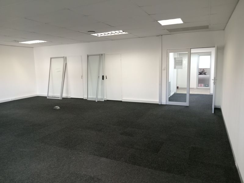 An open-plan office space featuring dark carpet tiles, a suspended ceiling with recessed lighting, and a glass partition door leading to an adjacent area.