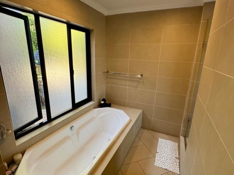 A clean, neutral-toned bathroom featuring a built-in bathtub, tiled walls and flooring, and a large frosted window for natural light.