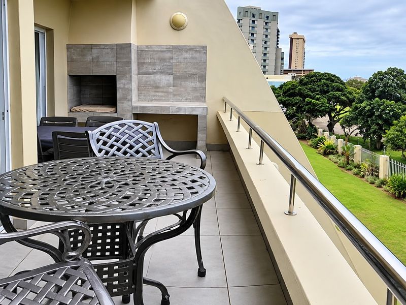 Spacious balcony featuring a built-in braai, tiled flooring, and a stainless steel balustrade, offering pleasant views of the surrounding greenery and coastline.