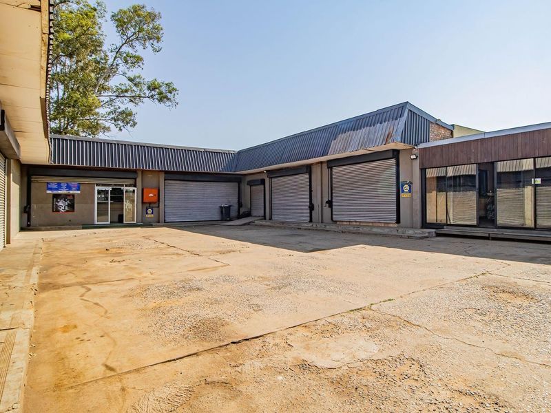 A commercial retail yard featuring multiple metal roller shutter doors, concrete paving, and glass storefronts, suitable for industrial or retail storage use.