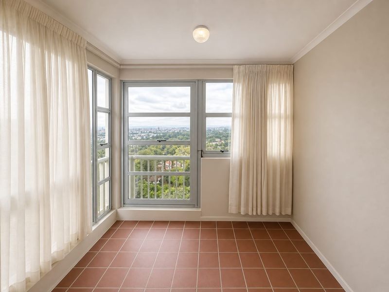 A bright, empty enclosed balcony featuring terracotta floor tiles, large aluminium-framed windows, and expansive views of the surrounding area.