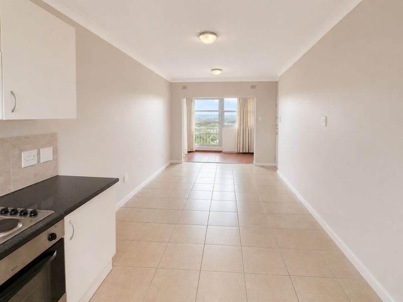 An open-plan kitchen and living area featuring tiled flooring, built-in oven and hob, leading to a balcony with scenic views.