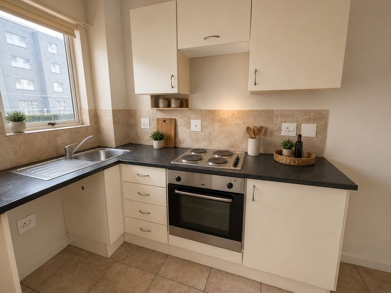A functional kitchen featuring cream cabinetry, dark laminate countertops, a built-in oven with electric hob, and tiled backsplash with neutral floor tiles.