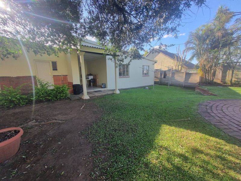Rear view of the property featuring a covered patio, paved area, and a spacious garden with established trees and perimeter walling.