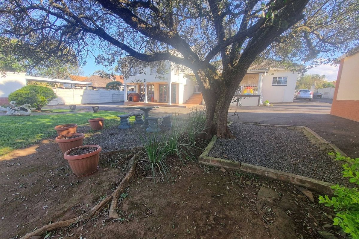 A spacious rear garden area featuring a large mature tree, paved driveway, and a covered patio attached to the main house structure.