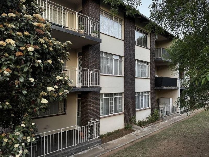 A multi-story apartment building exterior featuring brick accents, metal balcony railings, and large multi-pane windows overlooking a landscaped garden area.