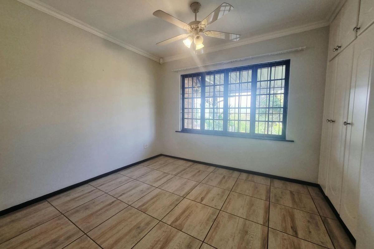 A bright, empty bedroom featuring wood-look tiled flooring, built-in cupboards, a ceiling fan, and a window fitted with security bars.