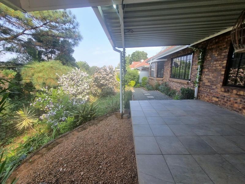 A covered outdoor patio area featuring tiled flooring and exposed brickwork, overlooking a landscaped garden and mature trees.