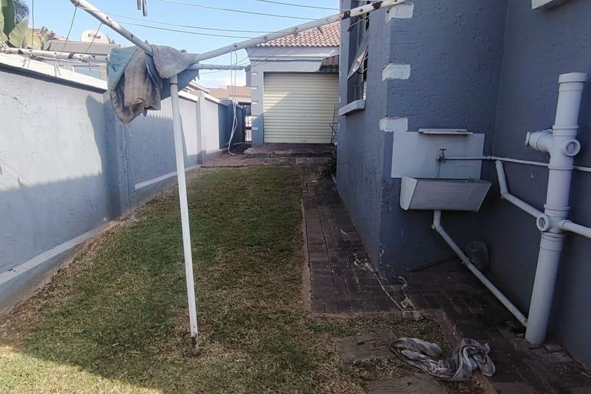 Rear exterior view showing a paved walkway, lawn area, external wash basin, and boundary wall leading towards a detached garage.