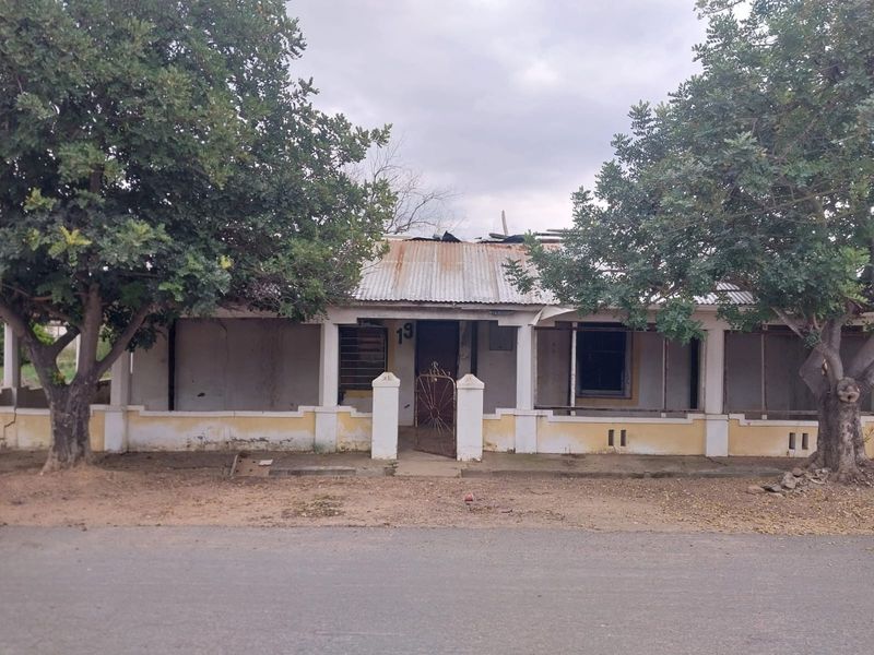 Front elevation of a single-story house featuring a covered veranda, corrugated metal roof, and masonry perimeter wall, showing significant signs of wear and structural deterioration.
