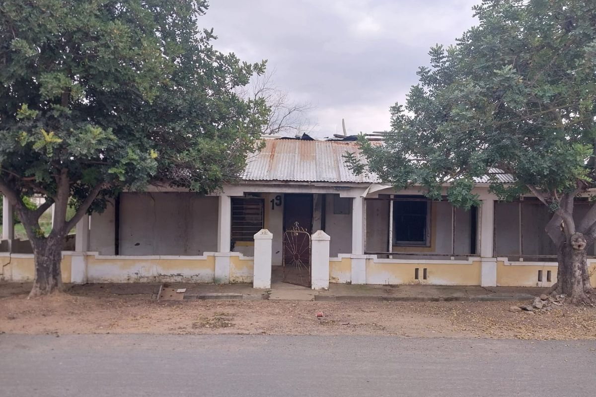 Front elevation of a single-story house featuring a covered veranda, corrugated metal roof, and masonry perimeter wall, showing significant signs of wear and structural deterioration.