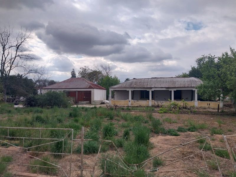 A view of a property featuring a main house with a covered veranda and a detached outbuilding, set on a large, overgrown plot.