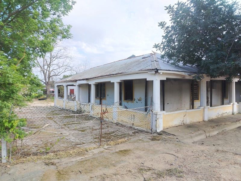 A weathered single-story house featuring a corrugated metal roof and a wrap-around veranda, situated on a large plot with existing fencing.