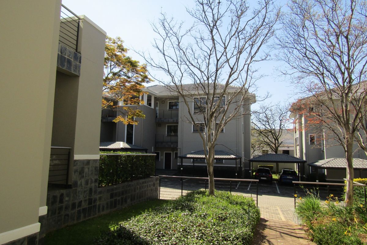 Exterior view of the apartment complex featuring paved driveways, covered parking bays, landscaped garden areas, and modern building facades with stone cladding.