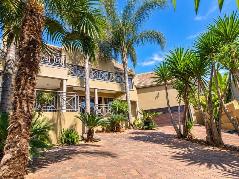Front elevation of a multi-story residence featuring a paved driveway, double garage, and upper-level balconies with metal railings, surrounded by established palm trees.