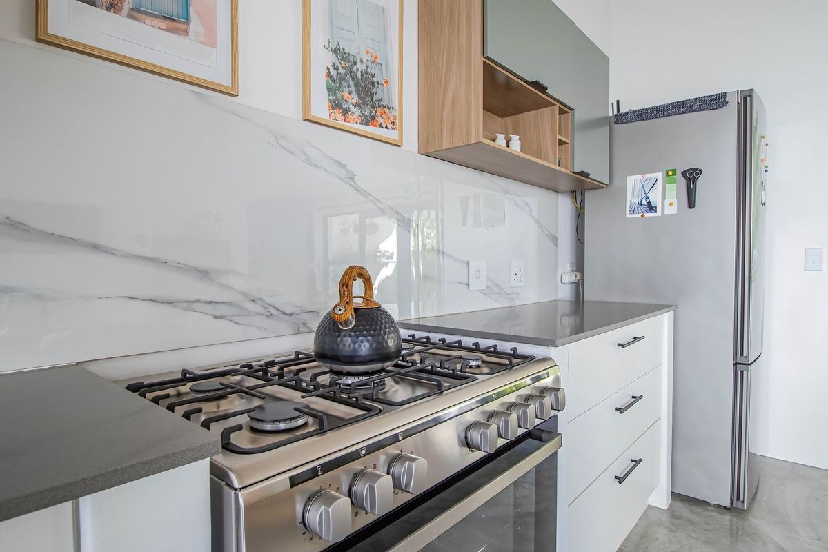 Modern kitchen featuring a stainless steel gas stove, marble-look splashback, stone countertops, and sleek white cabinetry with polished concrete flooring.