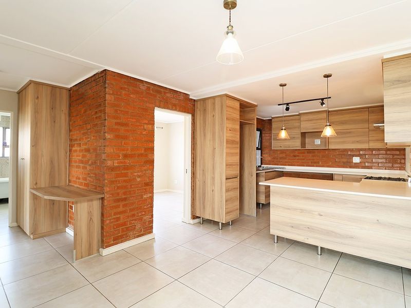 Modern open-plan kitchen and living area featuring exposed brick accents, light wood cabinetry, a central island, and neutral ceramic floor tiles.