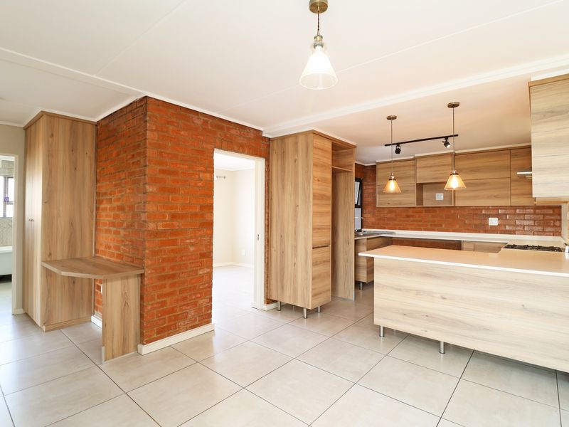 Modern open-plan kitchen and living area featuring exposed brick accents, light wood cabinetry, a functional kitchen island, and neutral ceramic floor tiles.