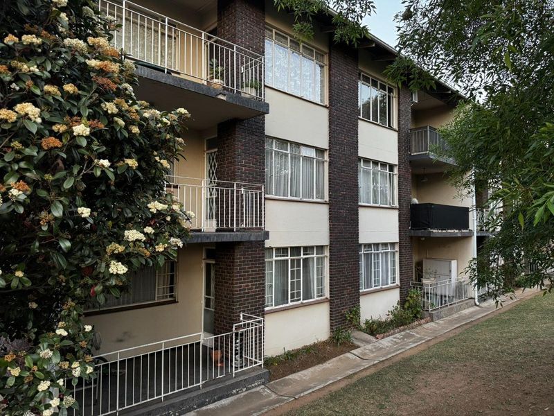 A multi-story apartment building exterior featuring brick accents, metal balcony railings, and large windows overlooking a landscaped garden area.