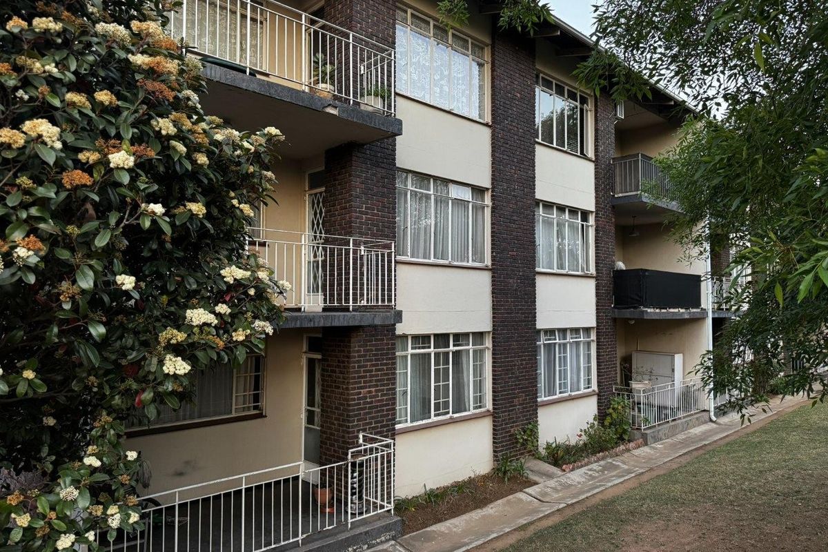A multi-story apartment building exterior featuring brick accents, metal balcony railings, and large windows overlooking a landscaped garden area.