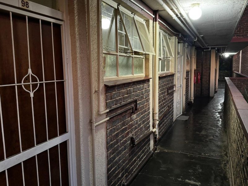 An exterior apartment corridor at night featuring exposed brickwork, security gates, and surface-mounted utility piping.