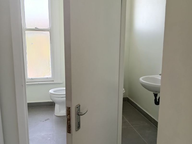 A functional bathroom featuring a toilet, wall-mounted basin, and tiled flooring, viewed through an open doorway.