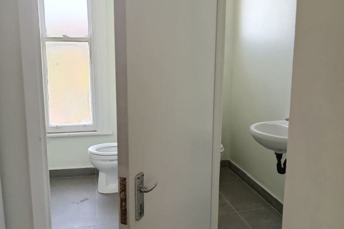 A functional bathroom featuring a toilet, wall-mounted basin, and tiled flooring, viewed through an open doorway.